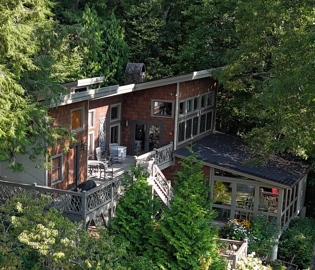 A wooden deck with decorative railing and large windows, surrounded by lush greenery. The inviting outdoor space encourages relaxation, blending the indoors with the natural environment.