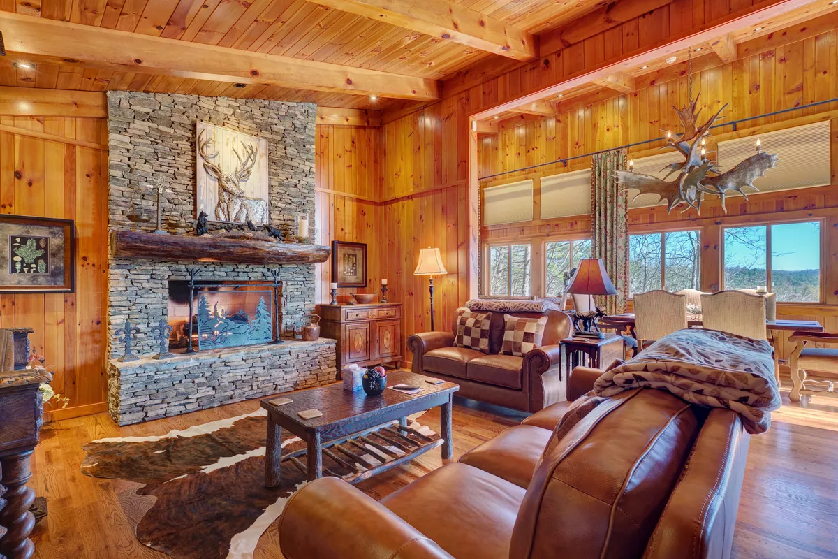 A warm, inviting living room featuring a prominent stone fireplace, comfortable buttery leather sofas, wooden walls, and large windows showcasing mountain views. The space is designed for both relaxation and entertaining, with natural light streaming in to highlight the cozy atmosphere.