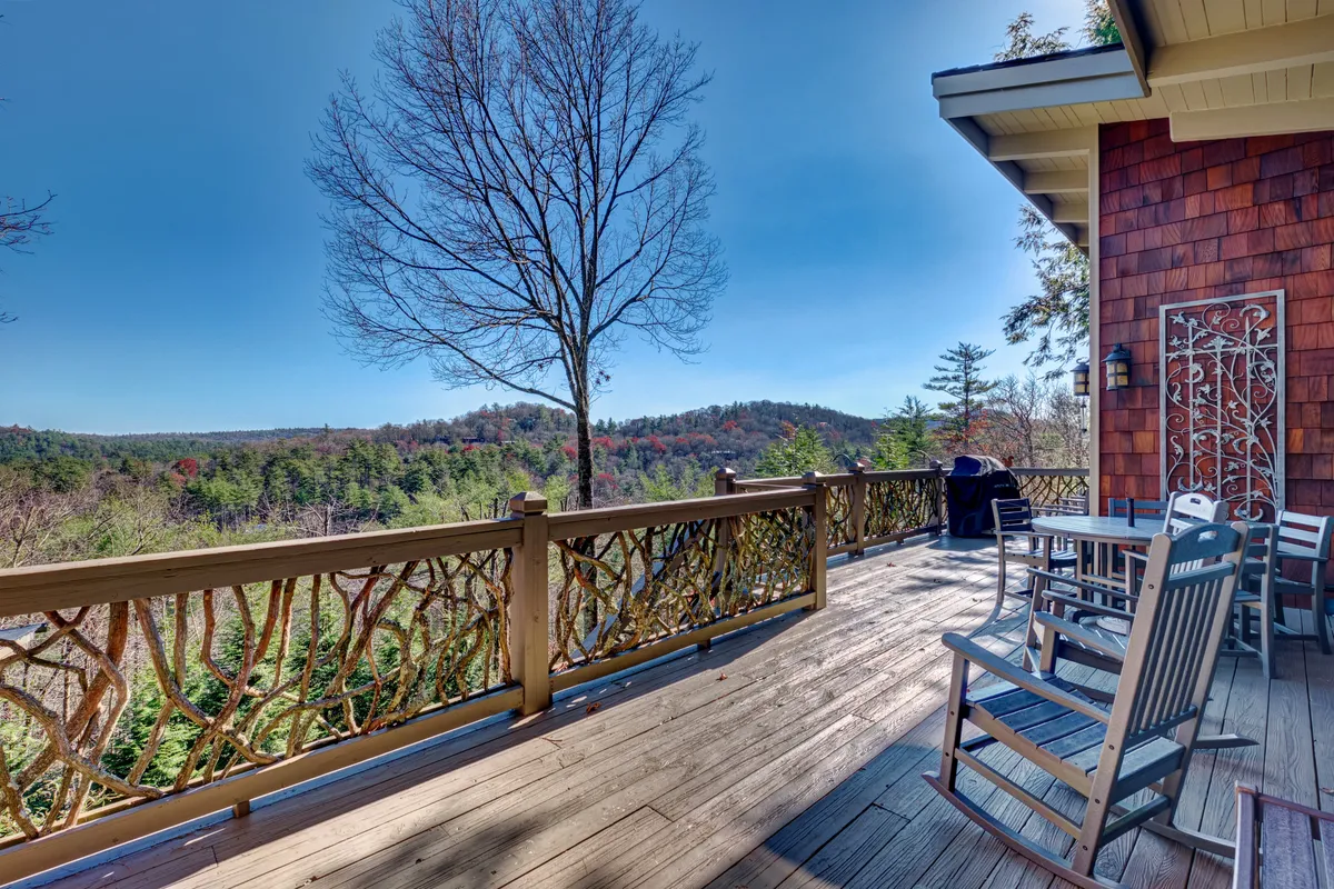 The image showcases a spacious wooden deck adorned with classic rocking chairs, set against a backdrop of sweeping mountain views. The decorative railing adds rustic charm, while the surrounding native trees and lush rhododendrons frame the scene, creating a sense of privacy and immersion in nature. The tranquil setting invites relaxation and outdoor gatherings, with ample space for enjoying sunrises, stargazing, or summer barbecues.