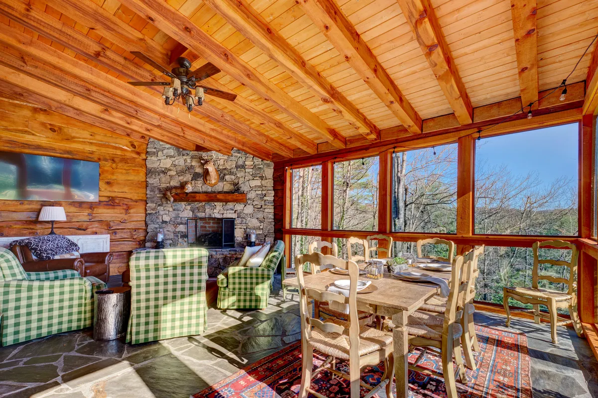 A sun-filled glass screen room featuring a robust stone fireplace, checkerboard lounge chairs, a spacious dining table with chairs, and expansive windows that frame beautiful mountain views. The setting balances comfort and elegance, with plenty of space for both dining and relaxation, as well as a flatscreen TV for entertainment.