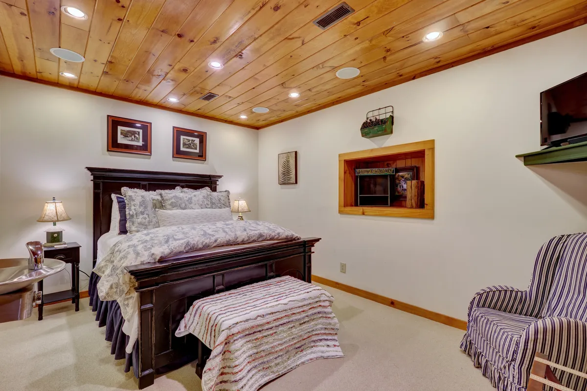 A warmly lit bedroom featuring a comfortable bed with decorative pillows, a nightstand with a lamp, a cozy striped chair, and tasteful artwork on the walls. The overall setting feels private and inviting, ideal for relaxation and peaceful rest.