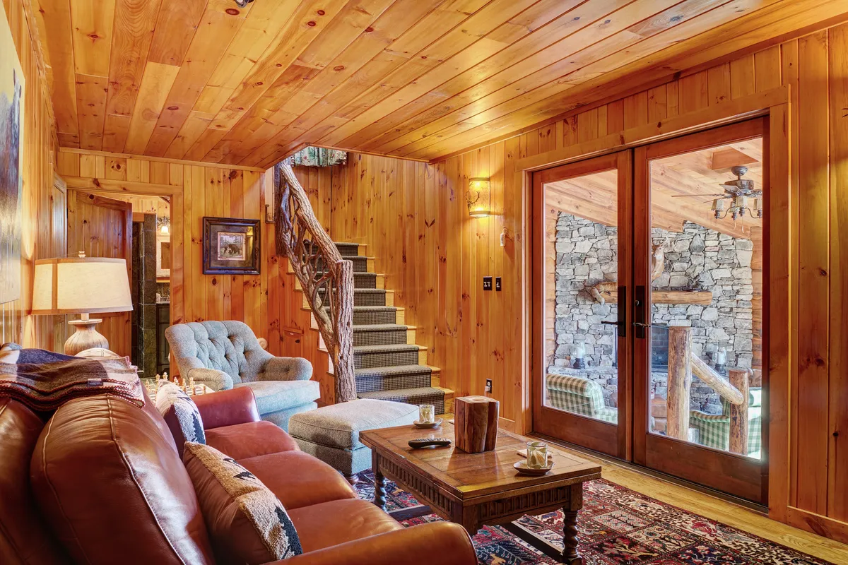 This interior image features a wooden staircase and a striking stone fireplace set against large windows. The windows allow abundant natural light to enter and offer glimpses of the surrounding scenery, subtly suggesting the home's connection to nature and the outdoors. The scene is inviting, with the promise of warmth and relaxation after a day outside.