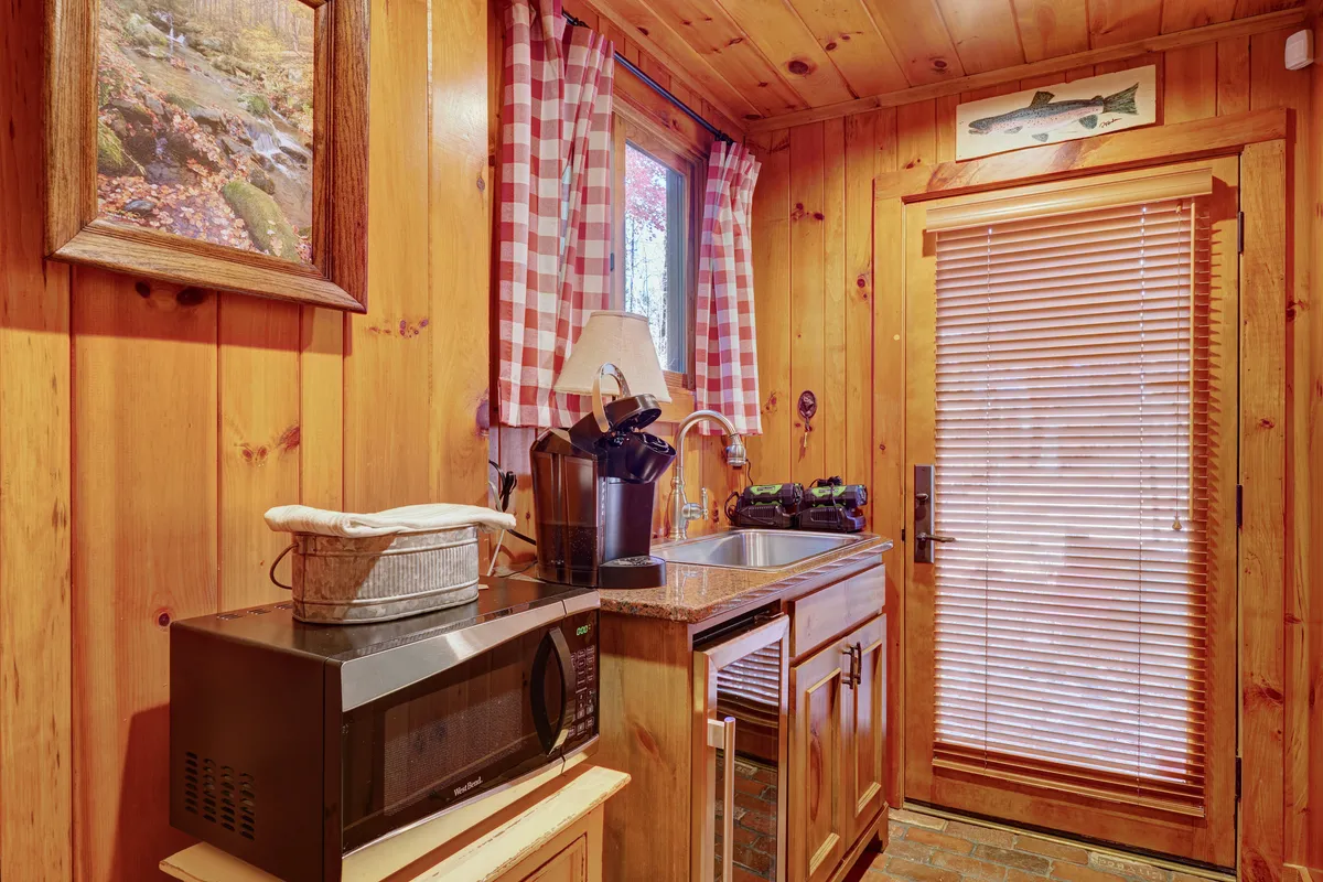 A welcoming kitchenette area featuring wooden cabinetry, a sink, microwave, and a window with charming checkered curtains. Framed nature artwork adds a touch of hominess. The space is perfectly suited for preparing quick meals or enjoying a morning coffee in private.