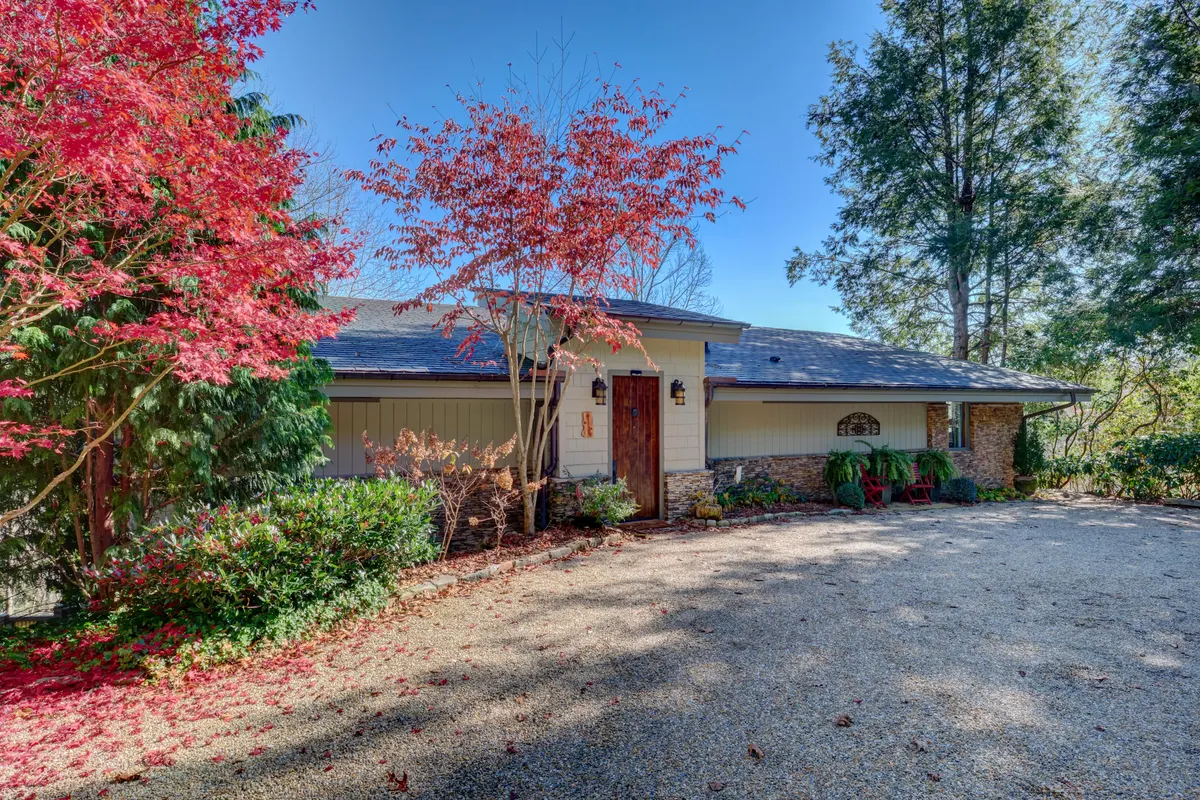 The image shows the inviting exterior of a mountain retreat, highlighted by a vibrant red tree, a charming stone-accented entryway, and a gravel driveway. The porch area suggests a welcoming spot for morning coffee or gathering outdoors, surrounded by natural beauty.