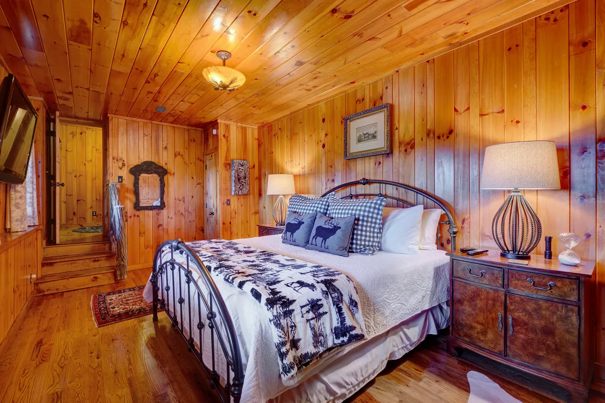 A tranquil bedroom with wooden walls, an inviting bed adorned with decorative pillows, a nightstand with lamps on both sides, and soft lighting that enhances the room’s comfort. The setting exudes peace and is ideal for unwinding after a long day.