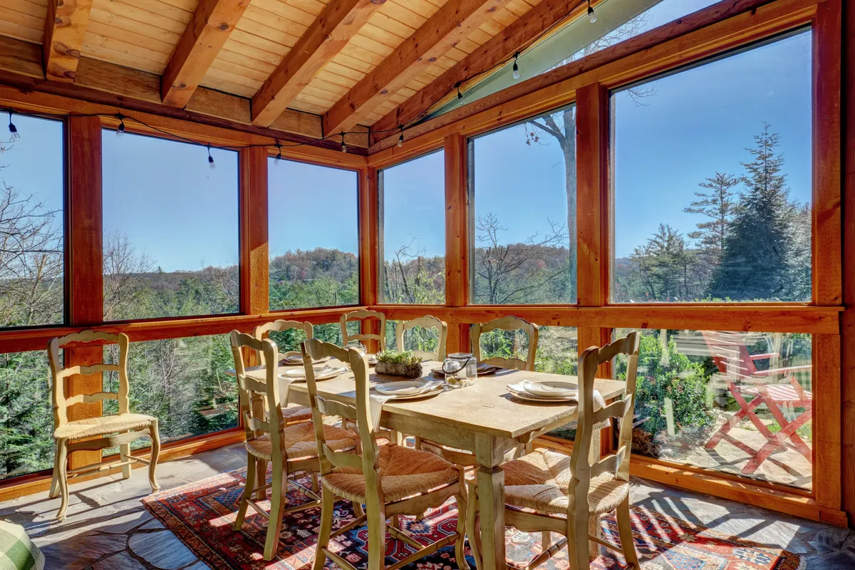 A warm, welcoming dining room with a large wooden table, surrounded by chairs and illuminated by sunlight streaming through oversize windows. Wooden beams and a cozy textile rug give the space a rustic charm, while the panoramic views outside hint at the home's beautiful natural surroundings.