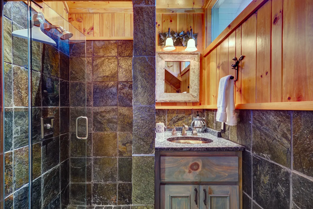 A cozy bathroom with a granite countertop, wooden cabinetry, a framed mirror, and a spacious, inviting shower area. The rustic decor and warm lighting create a relaxing atmosphere perfect for rejuvenation.